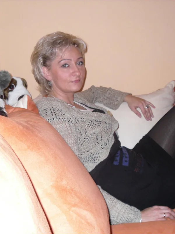 Femme aux cheveux blonds assise sur un canapé orange, entourée de peluches, portant un pull tricoté et une jupe noire.
