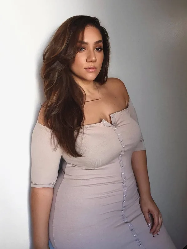 Femme aux cheveux bruns ondulés portant une robe beige ajustée, debout contre un mur clair, regardant l'objectif.