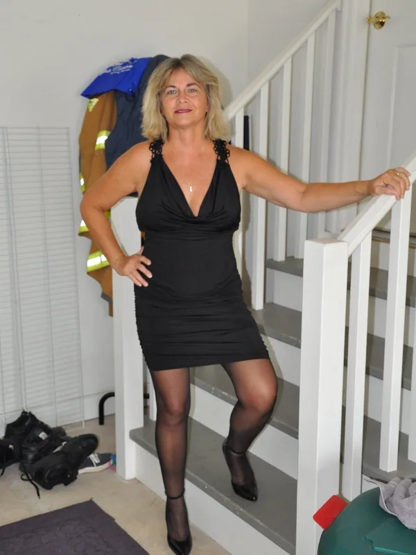 Femme blonde en robe noire posant sur un escalier intérieur, regardant l'objectif, dans un cadre domestique.