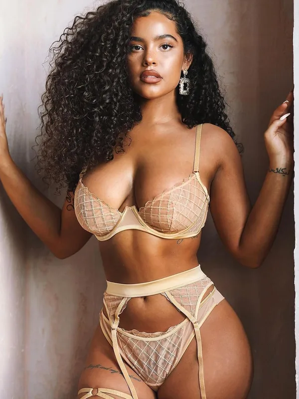 Femme aux cheveux bouclés portant une lingerie beige, appuyée contre un mur, regardant l'objectif.