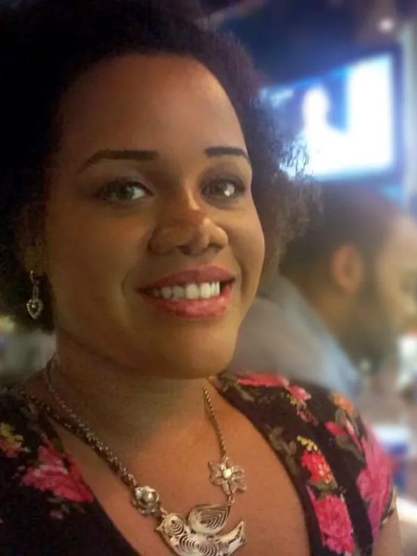 Portrait d'une femme souriante aux cheveux bouclés, portant un haut fleuri, tenant un verre dans un lieu animé la nuit.