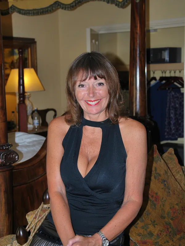 Femme assise sur un fauteuil dans une chambre, portant un haut noir et une jupe argentée, souriante devant un lit à baldaquin.