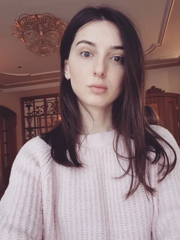 Portrait d'une femme aux cheveux bruns portant un pull rose pâle, regardant l'objectif dans un intérieur lumineux.