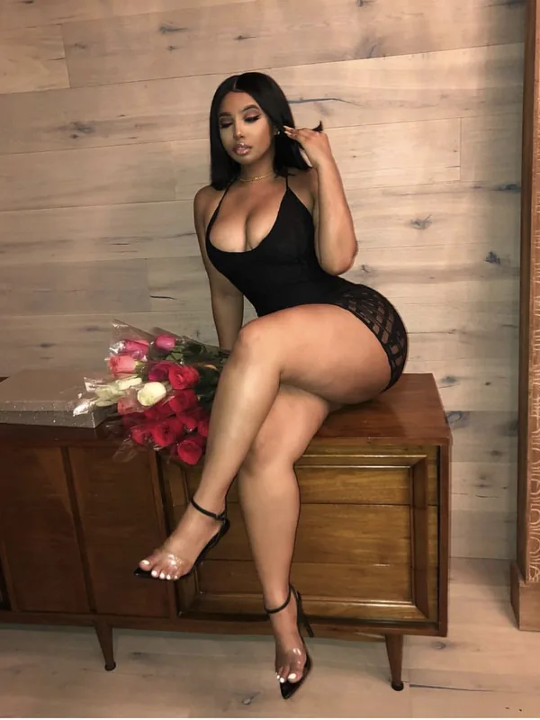 Femme aux cheveux noirs portant un body noir et des talons hauts, assise sur un meuble en bois avec un bouquet de roses à côté d'elle.