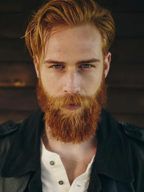 Portrait d'un homme aux cheveux et barbe roux, regardant l'objectif, vêtu d'une chemise blanche et d'une veste noire.