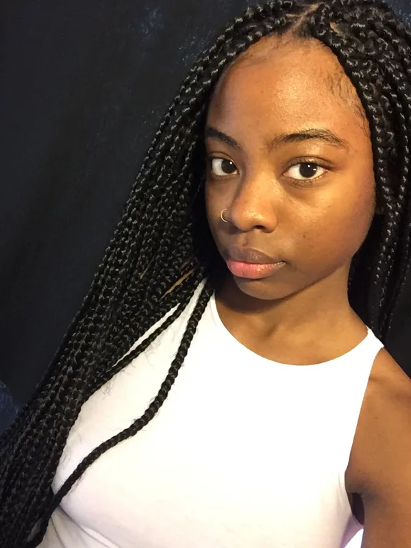 Portrait d'une jeune femme aux cheveux tressés, portant un haut blanc, regardant l'objectif devant un fond sombre.