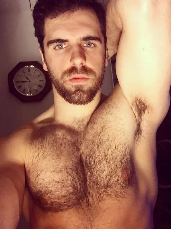 Homme torse nu regardant l'objectif, bras levé, dans une pièce éclairée par une lumière chaude, horloge en arrière-plan.