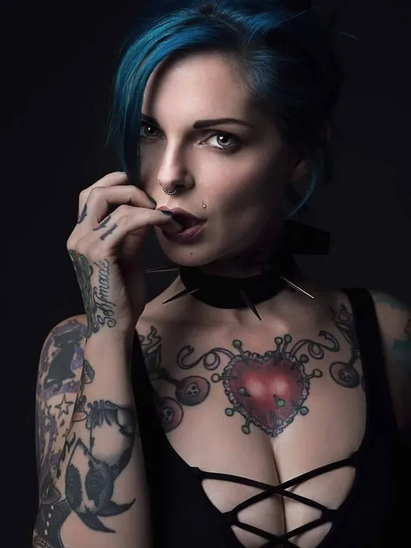 Portrait d'une femme aux cheveux bleus, tatouée, portant un haut noir à lacets et un collier à pointes, regardant l'objectif sur fond noir.