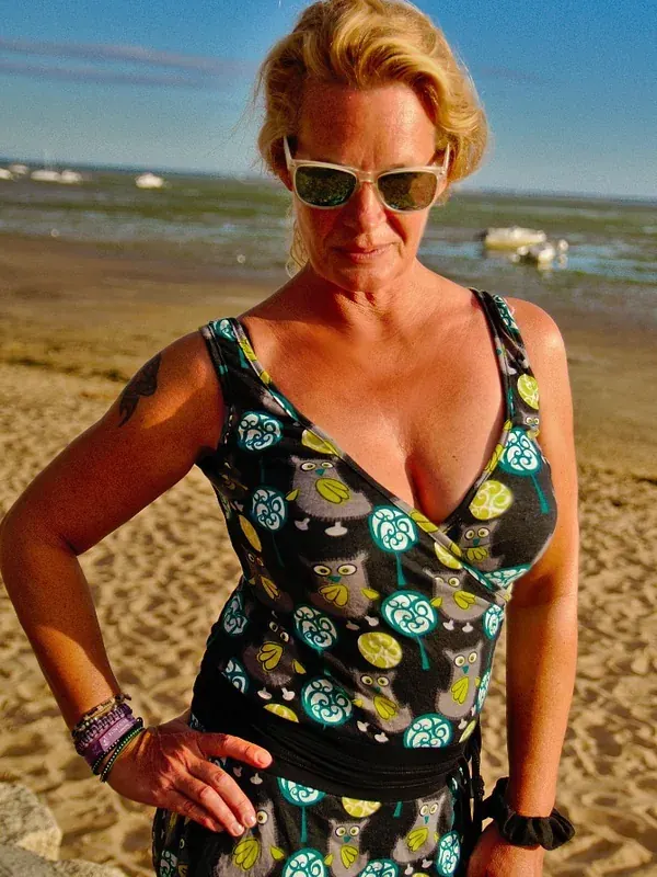 Femme blonde en robe à motifs d'hiboux sur une plage, portant des lunettes de soleil, regardant l'objectif.