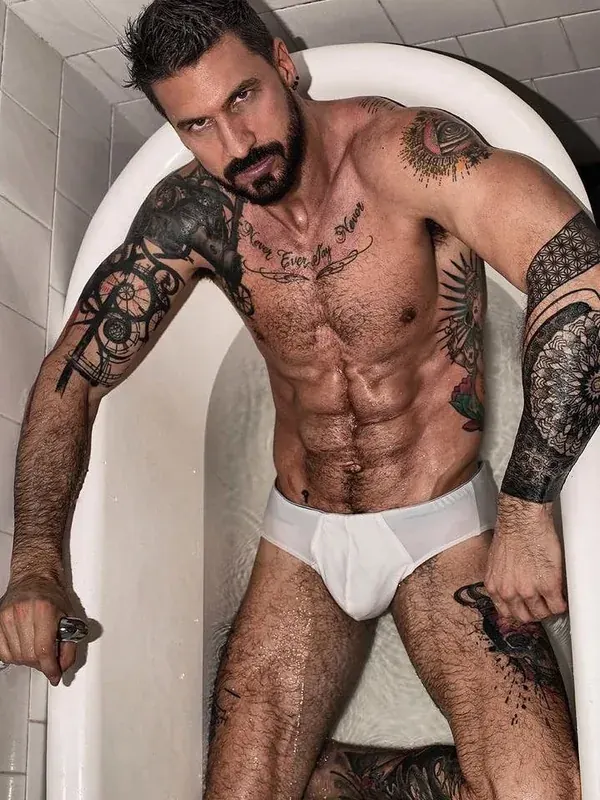Homme tatoué en sous-vêtement blanc assis dans une baignoire, regardant l'objectif.