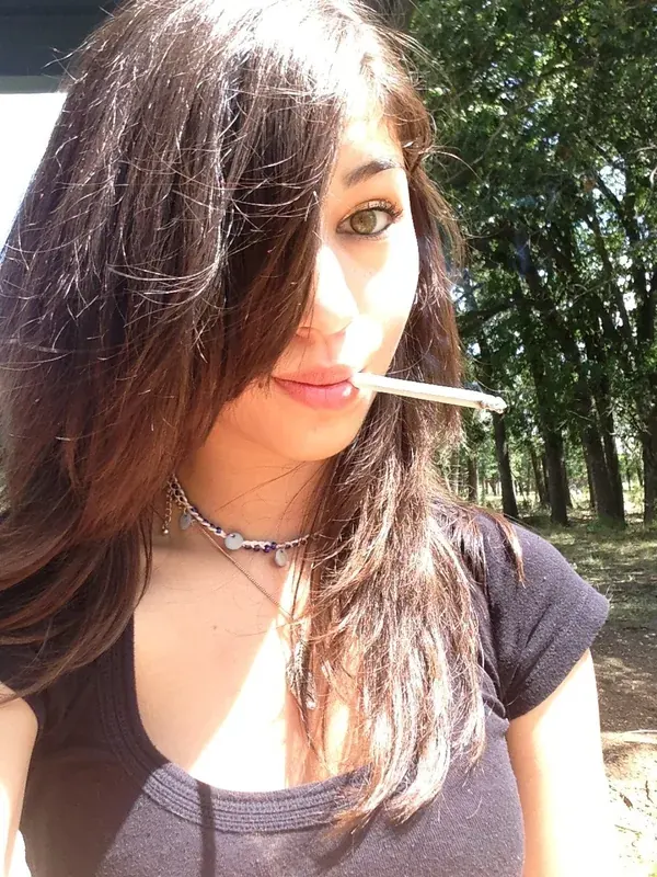 Portrait en gros plan d'une femme aux cheveux bruns tenant une cigarette, en extérieur sous un soleil vif, avec des arbres en arrière-plan.