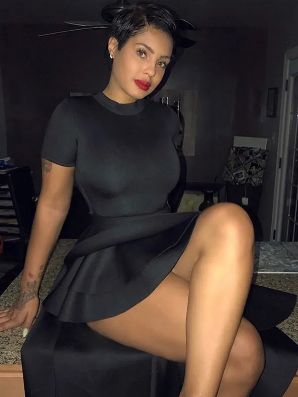 Femme aux cheveux courts portant une robe noire, assise sur une surface sombre, regardant l'objectif.