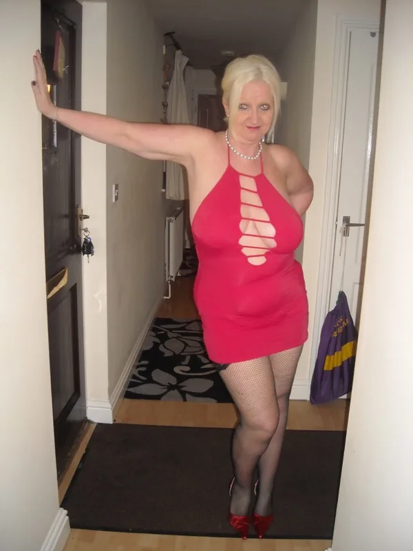 Femme blonde en robe rouge moulante avec découpes, collants à mailles et chaussures rouges, posant dans un couloir intérieur.
