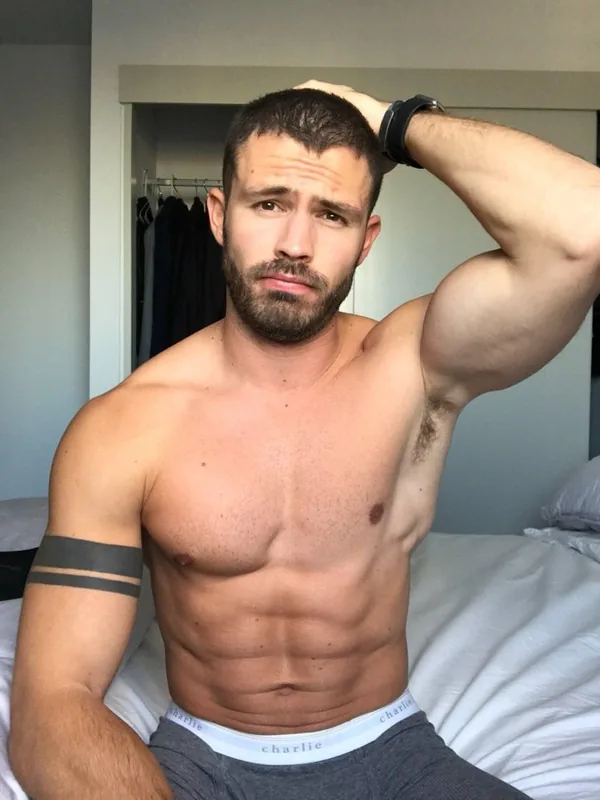 Homme torse nu assis sur un lit, regardant l'objectif, portant un short gris et un caleçon blanc.