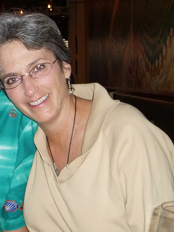 Portrait en buste d'une femme aux cheveux gris courts, portant des lunettes et un haut beige, souriant dans un cadre intérieur chaleureux.