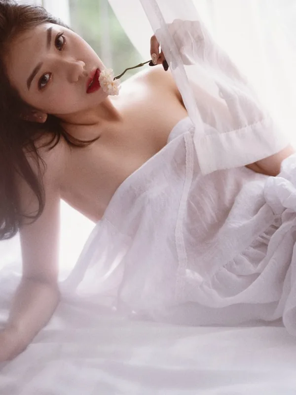 Femme aux cheveux bruns allongée sur un lit blanc, vêtue d'une chemise de nuit légère, tenant une fleur près de sa bouche.