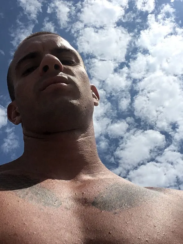 Portrait en contre-plongée d'un homme torse nu sous un ciel bleu nuageux, regardant l'objectif.