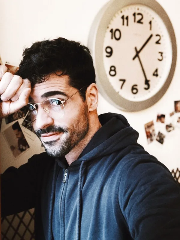 Portrait d'un homme aux cheveux foncés et lunettes, souriant légèrement, devant un mur avec une horloge et des photos.