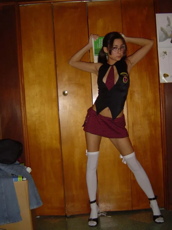 Femme en tenue d'écolière posant en pied devant une armoire en bois, tenant un livre derrière la tête.