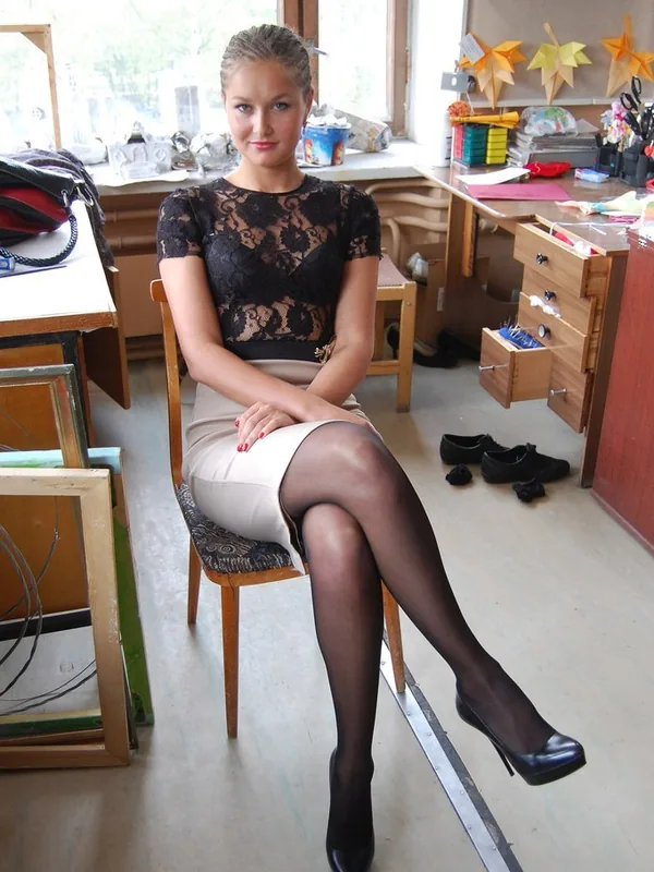 Femme assise sur une chaise en bois dans un atelier, portant une robe noire en dentelle et une jupe beige, regardant l'objectif.