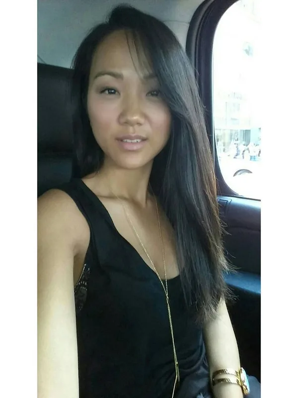 Portrait d'une femme assise dans une voiture, portant un haut noir et des bijoux dorés, regardant l'objectif.