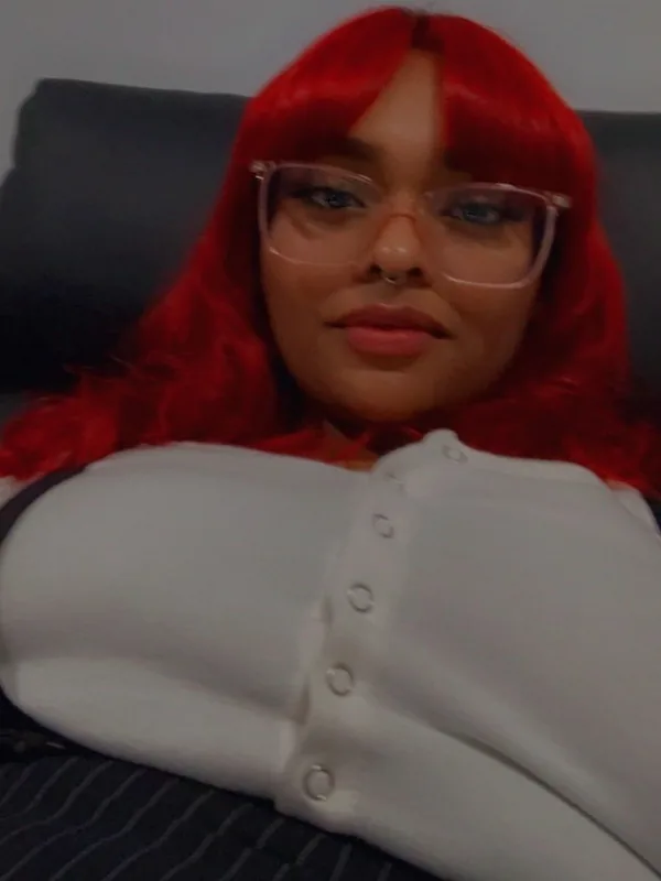 Portrait d'une femme aux cheveux rouges portant des lunettes et un haut blanc, allongée sur un canapé sombre.