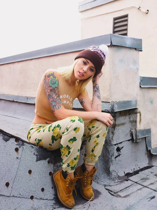 Femme blonde tatouée portant un bonnet, un haut beige et des pantalons floraux, assise sur un toit en béton.