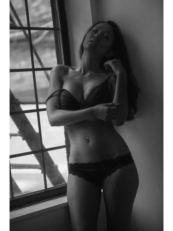 Femme en lingerie noire appuyée contre un mur près d'une fenêtre à carreaux, en noir et blanc.