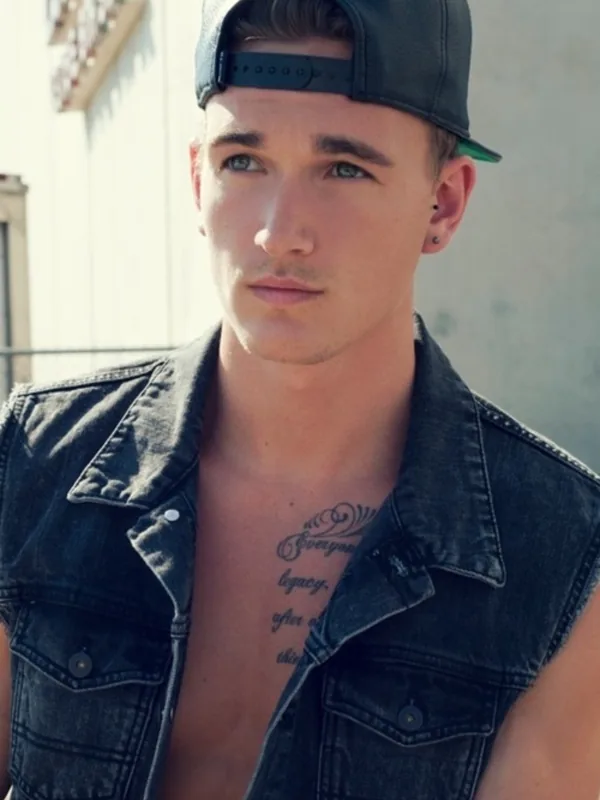 Portrait d'un jeune homme en gilet en jean noir, casquette à l'envers, regardant l'objectif, tatouage visible sur la poitrine.