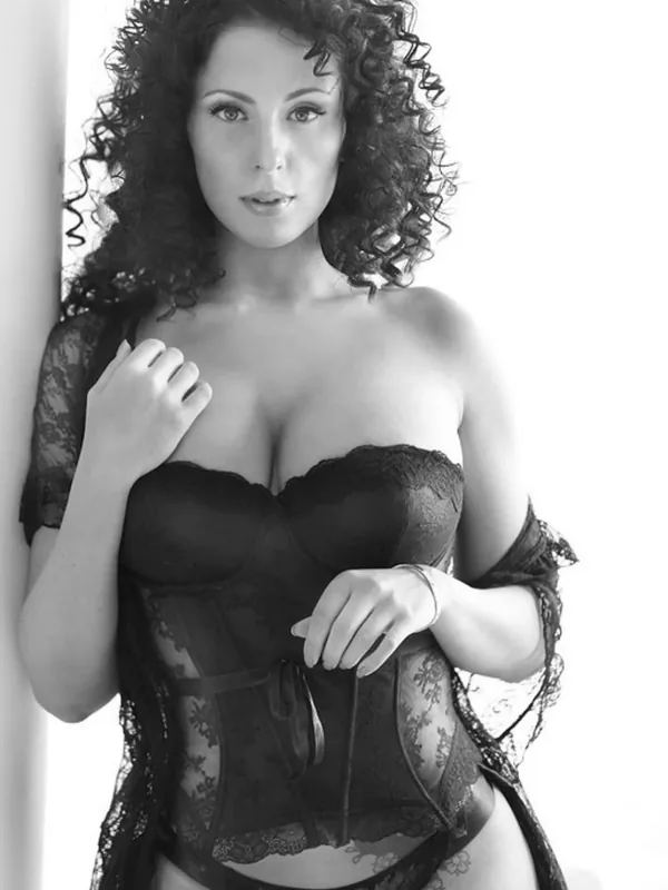 Portrait en noir et blanc d'une femme aux cheveux bouclés portant un corset et une cape en dentelle, appuyée contre un mur.