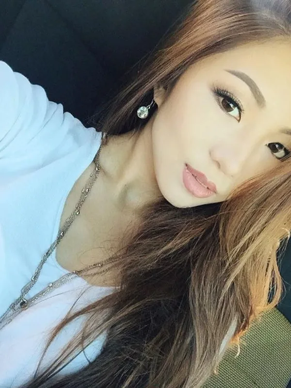 Portrait en selfie d'une femme aux cheveux bruns portant un t-shirt blanc et une chaîne, assise dans une voiture.