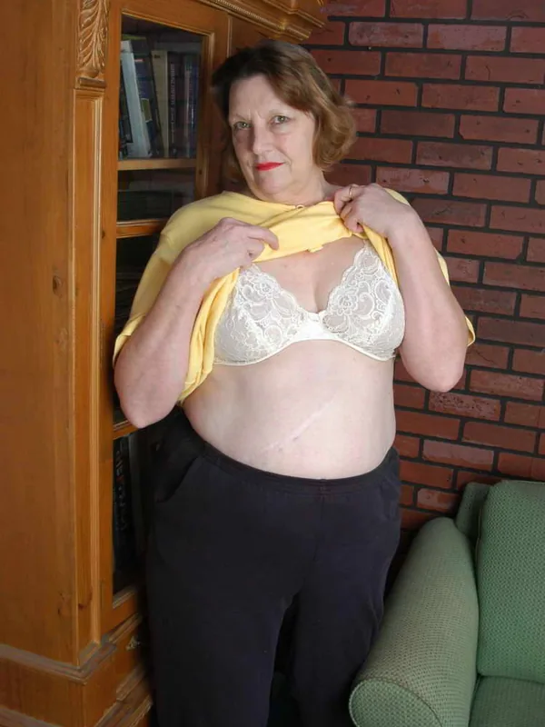 Femme aux cheveux bruns portant un soutien-gorge blanc et un haut jaune, debout devant un mur de briques et une bibliothèque.