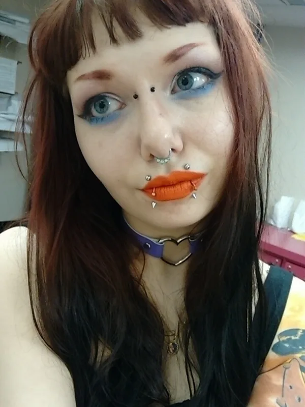 Portrait en gros plan d'une personne aux cheveux foncés, maquillage coloré, plusieurs piercings, collier violet et haut noir.