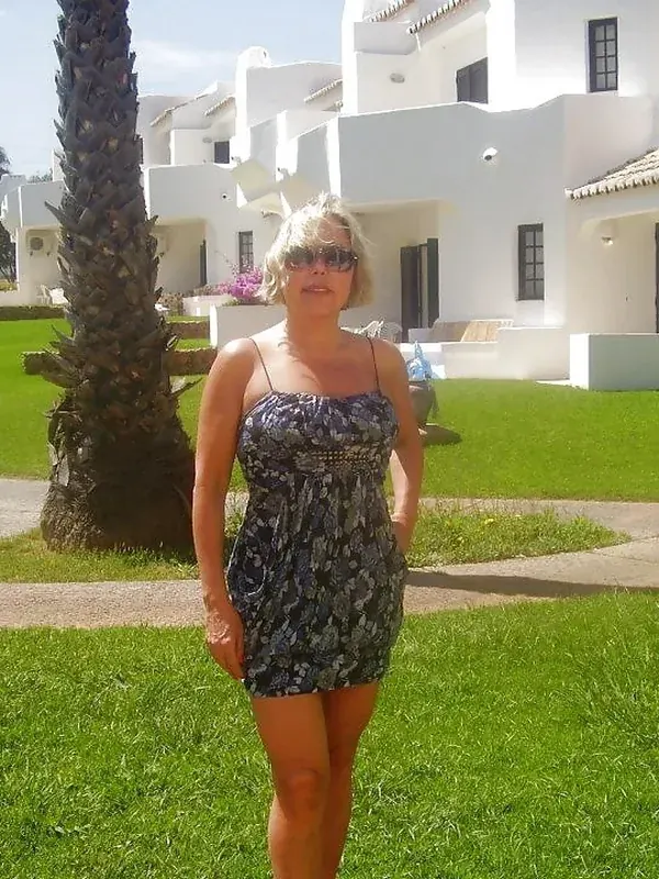 Femme blonde en robe courte posant sur une pelouse devant des bâtiments blancs et un palmier sous un ciel clair.
