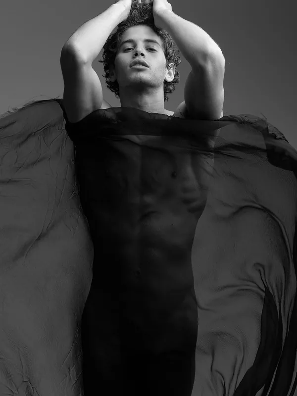 Portrait en noir et blanc d'un homme torse nu, les mains dans les cheveux, couvert partiellement d'un tissu noir translucide.