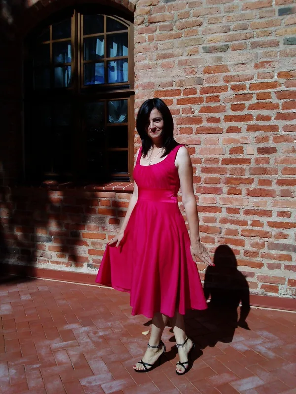 Femme en robe rose vif posant devant un mur en briques sous la lumière du soleil.