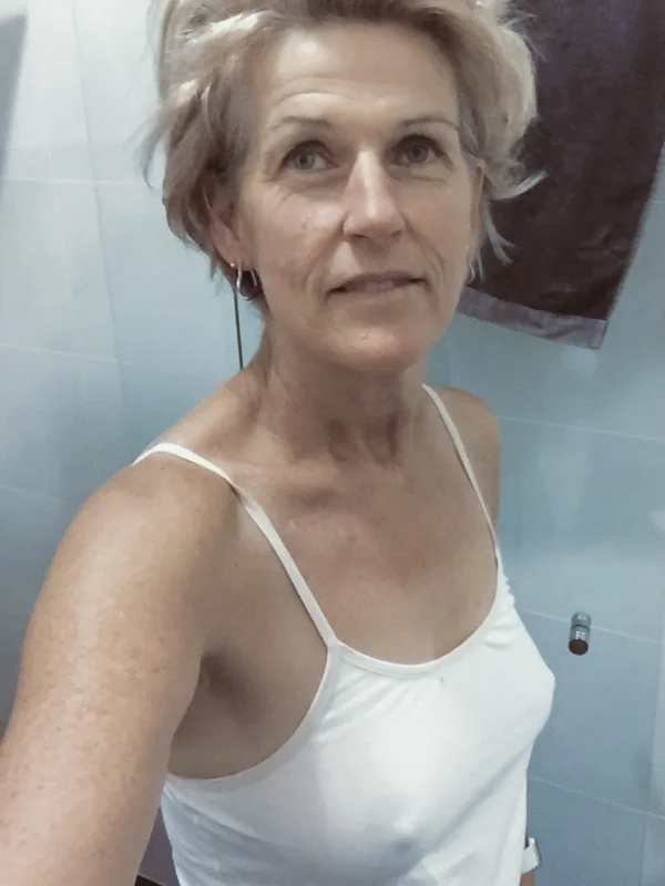 Portrait d'une femme aux cheveux courts blonds portant un débardeur blanc, prise en selfie dans une salle de bain.
