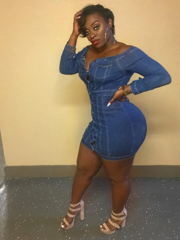 Femme debout contre un mur, portant une robe en jean bleu ajustée et des sandales à talons, regardant l'objectif.