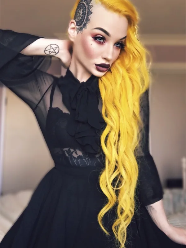 Femme aux cheveux jaunes vifs et tatouages, portant une robe noire transparente, regardant l'objectif.