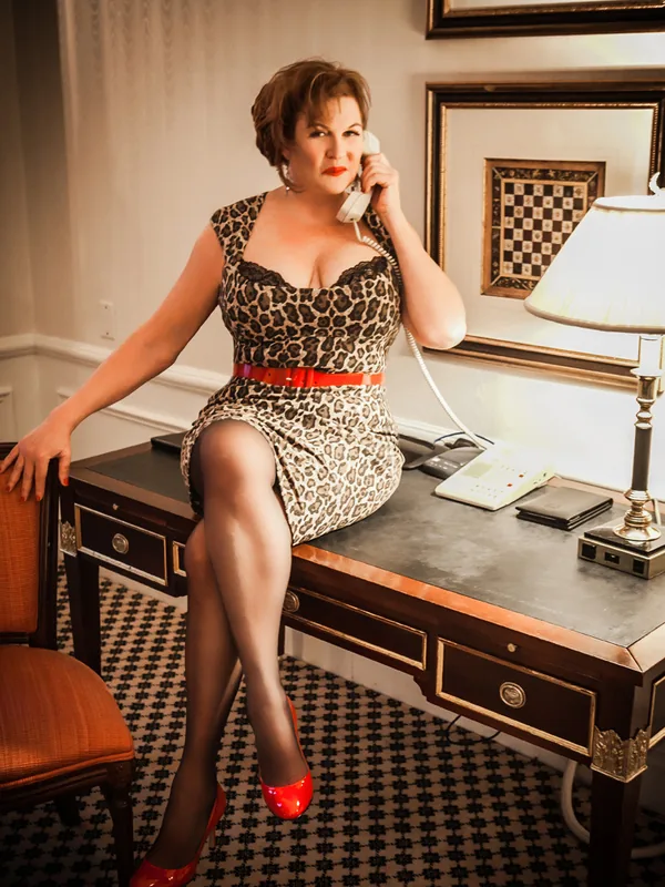 Femme assise sur un bureau, portant une robe léopard et des escarpins rouges, en train de parler au téléphone.