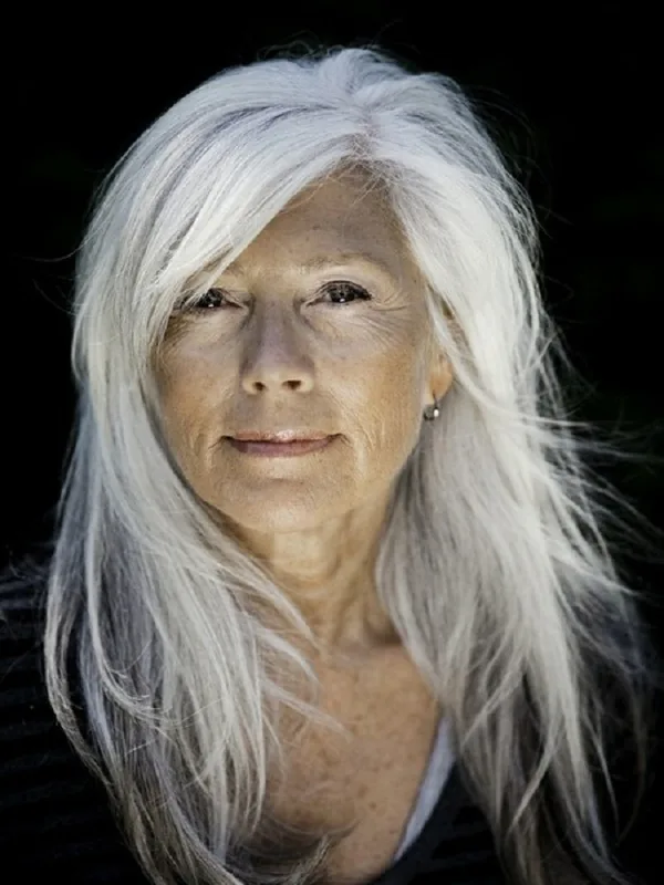 Portrait en buste d'une femme aux cheveux blancs longs, regardant l'objectif, sur fond noir.
