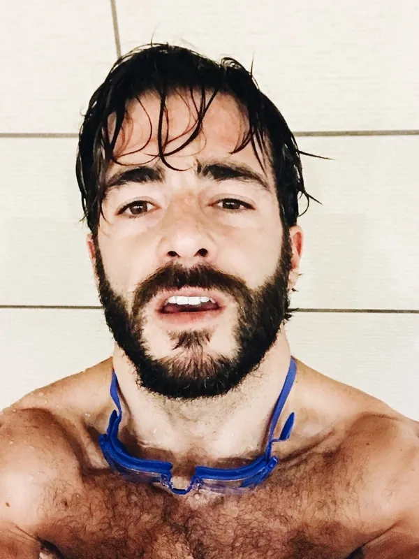 Homme torse nu avec barbe et cheveux mouillés, portant des lunettes de natation autour du cou, regardant l'objectif.