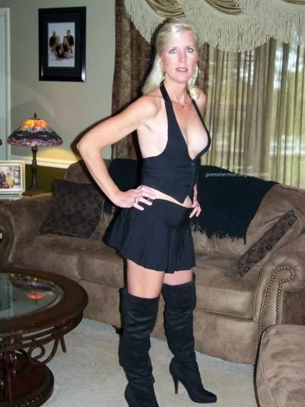 Femme blonde en robe noire et bottes hautes posant dans un salon avec canapé et lampe.