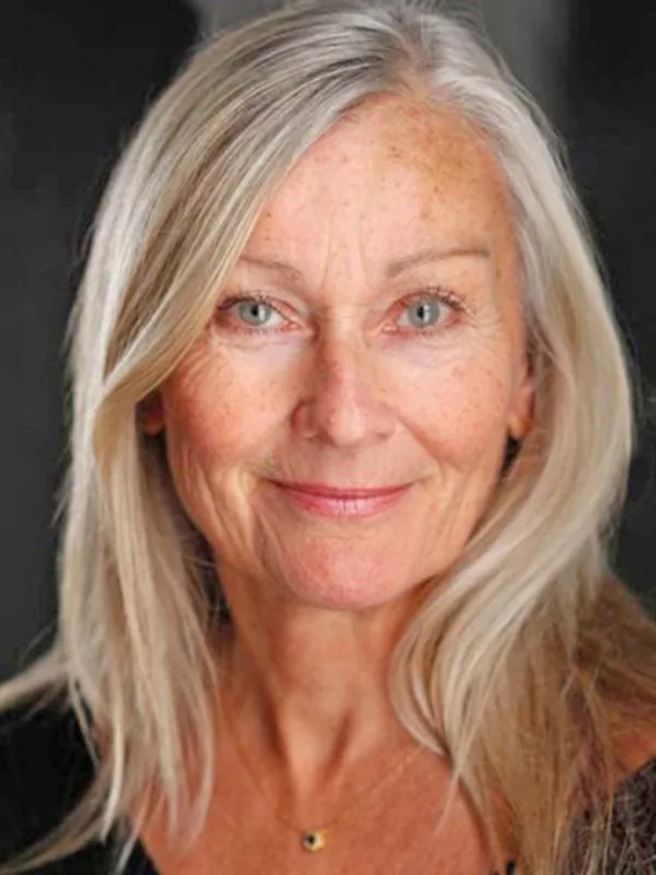 Portrait d'une femme aux cheveux blonds clairs, regardant l'objectif, souriante, sur fond sombre.