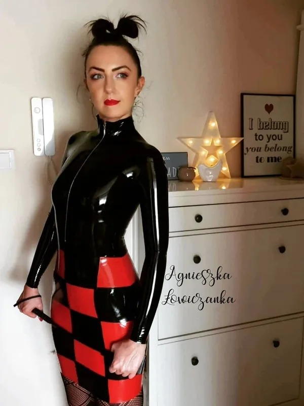 Femme en combinaison noire et rouge brillante tenant un fouet, posant près d'un meuble blanc avec une étoile lumineuse en arrière-plan.