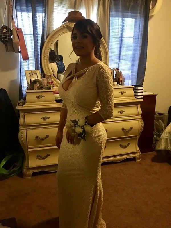 Femme en robe de mariée blanche tenant un bouquet, debout devant une commode avec miroir dans une chambre.