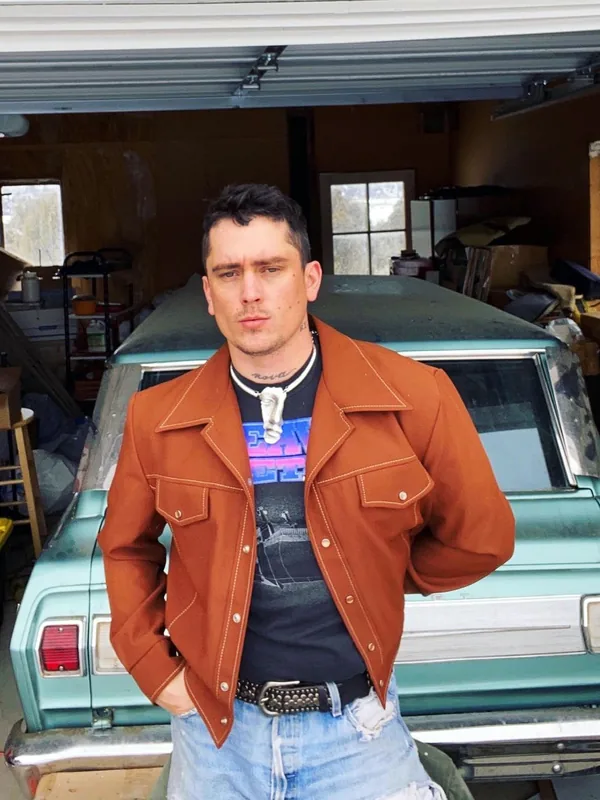 Un homme debout devant une voiture verte dans un garage, portant une veste en cuir marron et un jean déchiré.