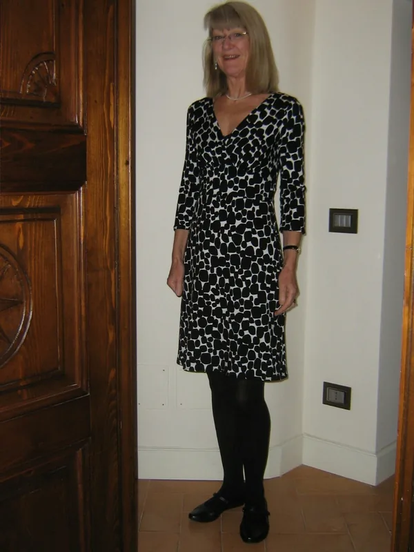 Femme debout en robe noire et blanche à motifs, collants noirs, chaussures noires, près d'une porte en bois dans un intérieur.
