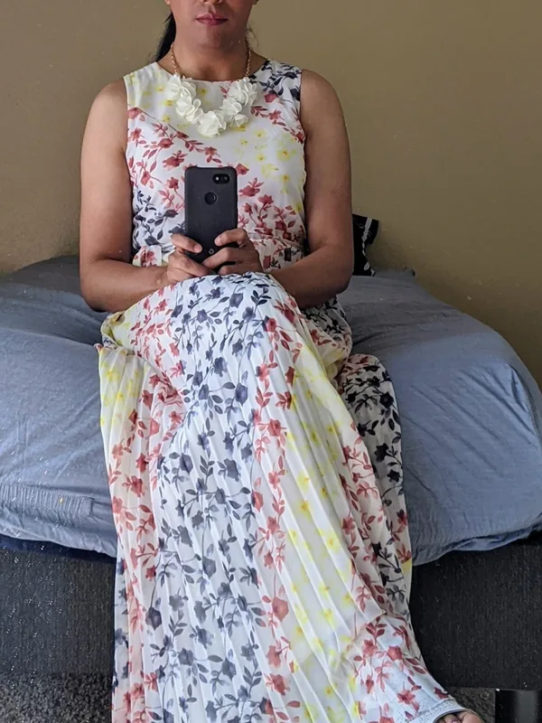 Femme assise sur un lit, portant une robe fleurie et des lunettes, tenant un téléphone devant elle.