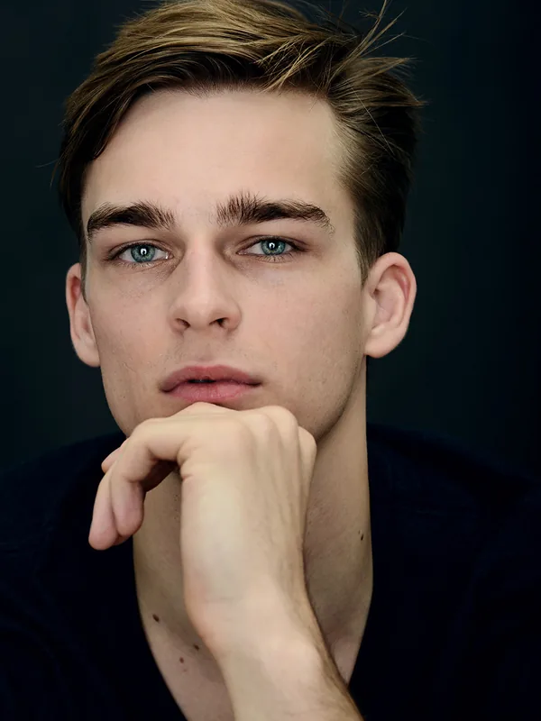 Portrait en gros plan d'un jeune homme aux yeux bleus, regardant l'objectif, main sous le menton, sur fond sombre.
